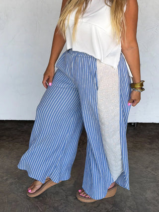 Woman wearing a white sleeveless top and blue striped pants, standing against a plain wall.
