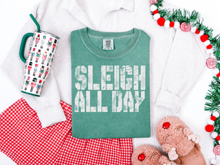 Green t-shirt with 'SLEIGH ALL DAY' text, mug with Christmas design, teddy bear, and Christmas tree on a white background