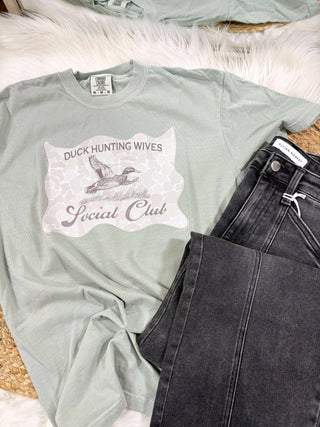 Light green t-shirt with graphic design and text, paired with black jeans on a textured surface.