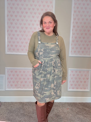 Person wearing a camouflage overall dress in a room with patterned wallpaper.
