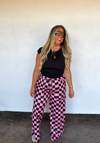 Woman wearing black top and pink checkered pants against a white wall