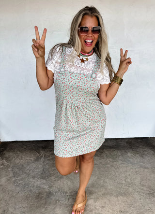 Woman wearing the Lilly Floral Skirt Overalls in a soft ribbed floral fabric. The skirt features oversized side pockets, a front bib pocket, and adjustable button straps. Styled with a fitted top underneath, perfect for a casual summer look.