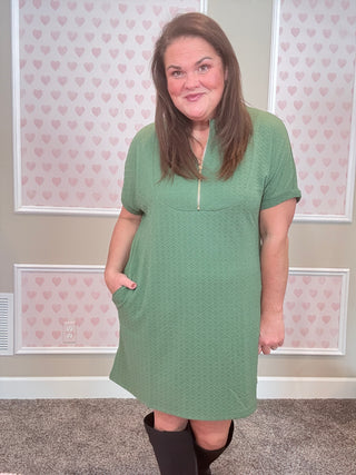 Woman wearing a green dress standing in a room with pink heart-patterned wallpaper.