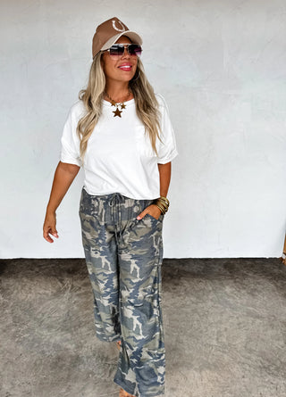 Woman wearing a white t-shirt, camouflage pants, and a cap against a plain background