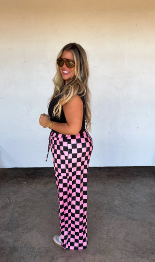 Woman wearing black top and pink checkered pants against a white wall