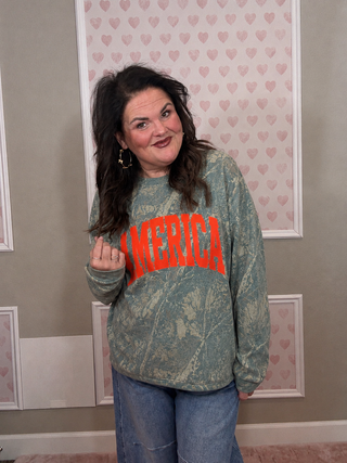 Person wearing a green sweatshirt with 'AMERICA' printed in red, standing in a room with heart-patterned wallpaper.