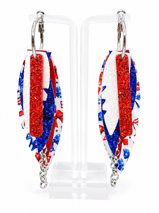 A pair of patriotic-themed dangle earrings displayed on clear acrylic stands. The earrings feature a distressed American flag pattern with red glitter, white glitter, and blue glitter sections, complete with silver-tone hook backings and dangling chain links.