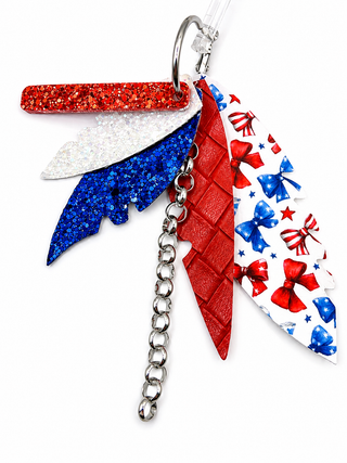 A patriotic-themed earrings featuring three stylized wings: a red glitter wing, a white iridescent wing, and a blue glitter wing, alongside a red quilted wing and a white wing printed with red bows and blue stars. The keychain is finished with a silver metal ring and a dangling silver chain.