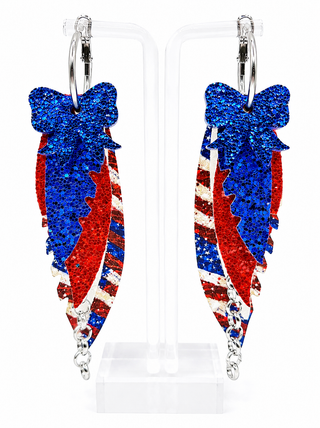 A pair of patriotic hoop earrings with dangling glittery bow and feather designs in red, white, and blue, displayed on clear acrylic stands against a white background.