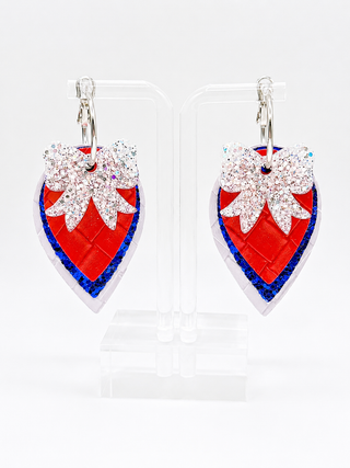 A pair of heart-shaped drop earrings displayed on clear acrylic stands against a white background. The earrings feature a textured red heart body with a blue glitter border, a large white glitter bow on top, and silver-tone hooks.