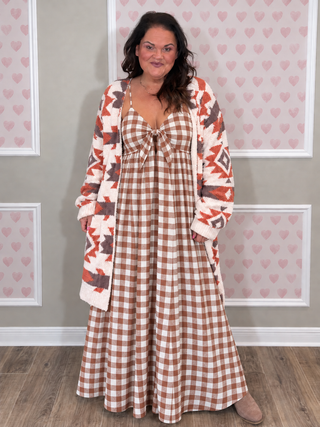 A woman modeling a long-sleeve maxi dress with a brown and white gingham pattern, featuring a tie at the neckline and a matching patterned cardigan draped over her shoulders, set against a decorative wall with pink hearts.
