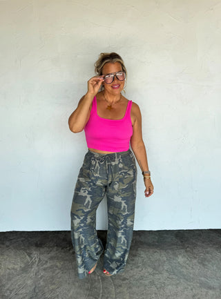 Woman wearing a pink top and camouflage pants against a white wall.