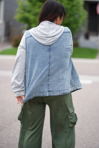 Person wearing a denim jacket over a light gray hoodie with green jumpsuit outdoors.