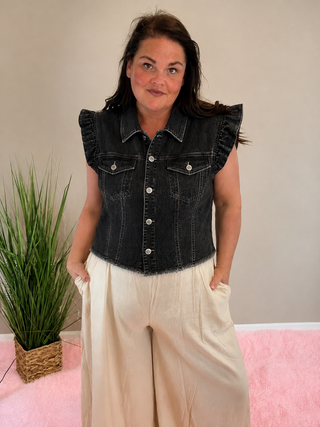 Woman wearing a faded black ruffle denim vest with cream wide-leg pants in a soft neutral indoor setting.