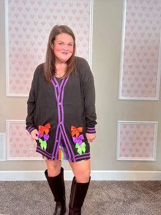 Woman wearing a black CARDIGAN with colorful bow accents in a room with heart-patterned wallpaper.