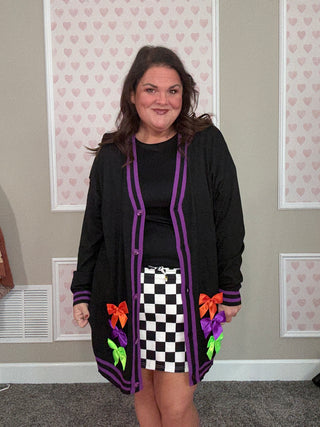 Person wearing a black cardigan with colorful bows in a room with patterned wallpaper.
