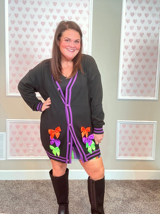 Woman wearing a black cardigan with purple trim and colorful bows in front of a wall with heart patterns.