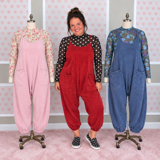 A woman stands between two mannequins wearing corduroy overall-style jumpsuits in pink and blue, each layered over a floral long-sleeve top. The woman wears a black-and-white polka dot top and black-and-white slip-on shoes.
