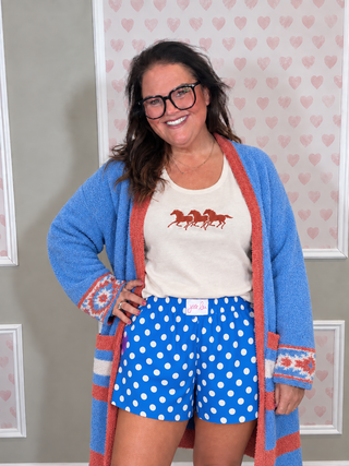 A woman wearing a blue knit cardigan with coral trim, a cream graphic t-shirt with red horses, and blue polka-dot shorts, standing in front of a heart-patterned backdrop.