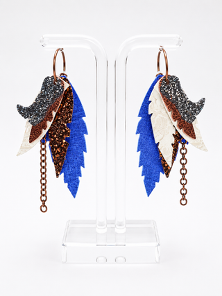 A pair of dangling feather earrings displayed on a clear acrylic stand against a white background. The earrings feature layered feather-like pieces in bright blue, white, and glittery brown, with a dark glittery black piece on each, hanging from copper-toned rings with short dangling chains.