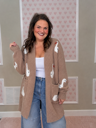 A woman smiling and wearing a beige knit cardigan with white goose motifs, paired with a white tank top and blue jeans, standing in front of a wall with pink heart wallpaper.