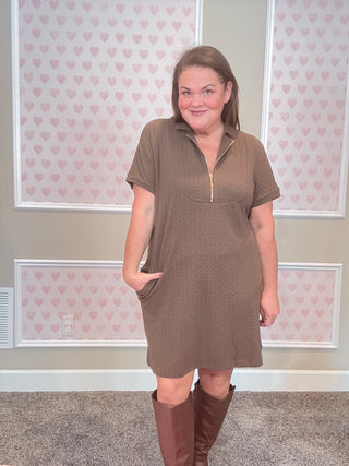 Woman wearing a brown dress with a zipper in front, standing against a wall with heart patterns.