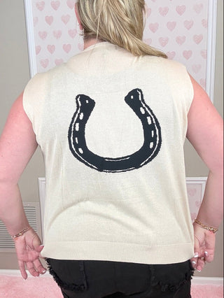Person wearing a cream-colored sleeveless top with a black horseshoe design on the back, standing in a room with heart-patterned wallpaper.