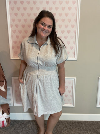 Woman wearing a light gray dress with a patterned border in a room with heart-shaped wallpaper.