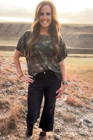 Woman in camouflage shirt and black pants standing in a field with mountains in the background
