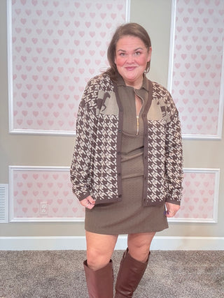 Woman wearing a patterned jacket over a dress with knee-high boots in a room with heart-patterned wallpaper.