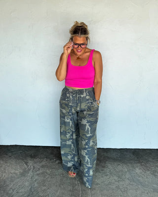 Woman wearing a pink crop top and camouflage pants against a white wall.