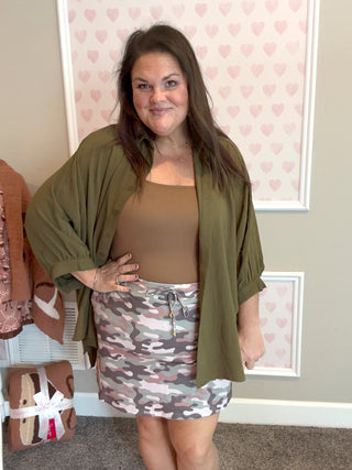 Woman wearing a green top, brown top, and camouflage skirt in a room with heart-patterned wallpaper.
