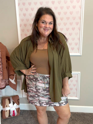 Woman wearing a green top, brown top, and camouflage skirt in a room with heart-patterned wallpaper.