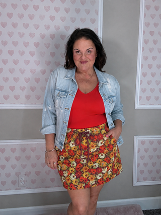 A woman modeling a casual outfit consisting of a light blue denim jacket, a bright red scoop-neck top, and a short floral mini skirt with orange, yellow, and white flowers, standing against a gray wall with pink heart-patterned wallpaper.