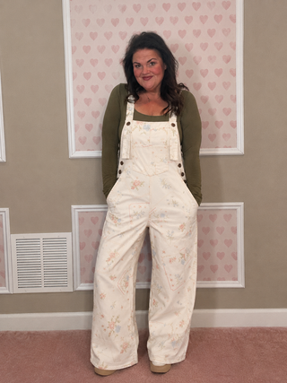 Woman wearing floral overalls and a green shirt in a room with heart-patterned wallpaper.