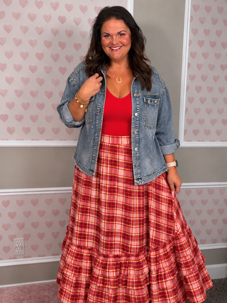 A woman modeling a red and pink plaid maxi skirt with a red top and a light blue denim jacket, standing in front of a wall with pink heart wallpaper.