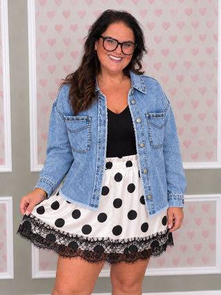 A woman wearing a light blue denim jacket over a black top and a white skirt with black polka dots and a black lace hem, standing in front of a pink heart-patterned wall.