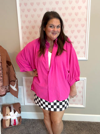 Woman wearing a bright pink shirt and checkered skirt in a room with heart-patterned wallpaper.