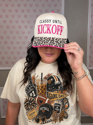 A woman wearing a cream trucker hat with a leopard print brim and pink trim, featuring the text 'CLASSY UNTIL KICKOFF' in black and pink. She is also wearing a graphic t-shirt with football and sports themes.