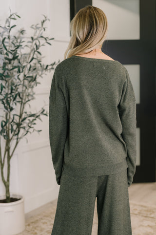 Person wearing a green sweater and pants in a room with a plant and framed picture.