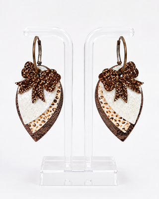 A pair of dangling earrings displayed on clear acrylic stands against a white background. Each earring has a brown glitter bow on a white textured heart-shaped drop with a brown leopard-print strip and brown faux-leather trim.
