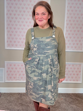 Person wearing a camo overall dress, standing in a room with pink heart-patterned wallpaper.