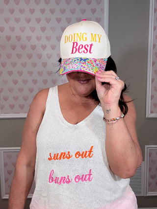 Person wearing a cap with 'Doing My Best' text and a tank top with 'suns out, buns out' text, against a heart-patterned wall.