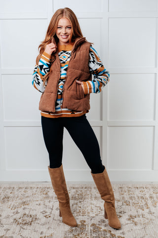 Woman wearing a patterned sweater, brown vest, black leggings, and brown boots against a white wall.