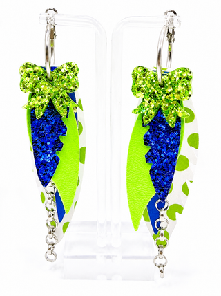 A pair of dangling earrings displayed on clear plastic stands against a white background. Each earring features a bright green glitter bow, a blue glitter center, a lime green textured side panel, a white side panel with green spots, and silver-tone hooks and chain drops.