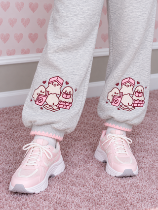 A person wearing light heather gray jogger sweatpants with pink and white cartoon ghost illustrations on the lower legs, paired with pink sneakers, standing on a pink carpet.