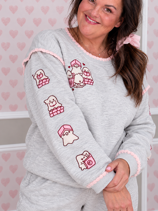 A woman wearing a light gray heathered pajama sweatshirt with pink ruffle trim and cute ghost-themed graphics, paired with matching gray pants against a pink heart-patterned background.