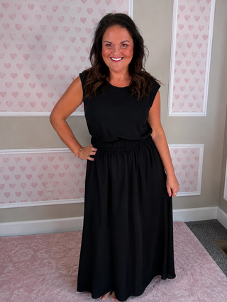 A woman wearing a sleeveless black maxi dress with a gathered elastic waist, standing in a room with pink heart-patterned wallpaper and a pink rug.