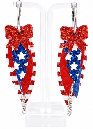 A pair of patriotic-themed dangle earrings displayed on a clear acrylic stand. The earrings feature a red glitter bow at the top, red and white glitter stripes, blue glitter chevron patterns, and white glitter stars, with silver-tone metal hooks and chains.