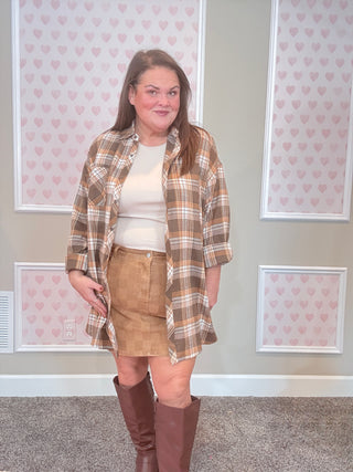 Woman wearing a plaid flannel, white top, tan skirt, and brown knee-high boots in front of a wall with heart patterns.